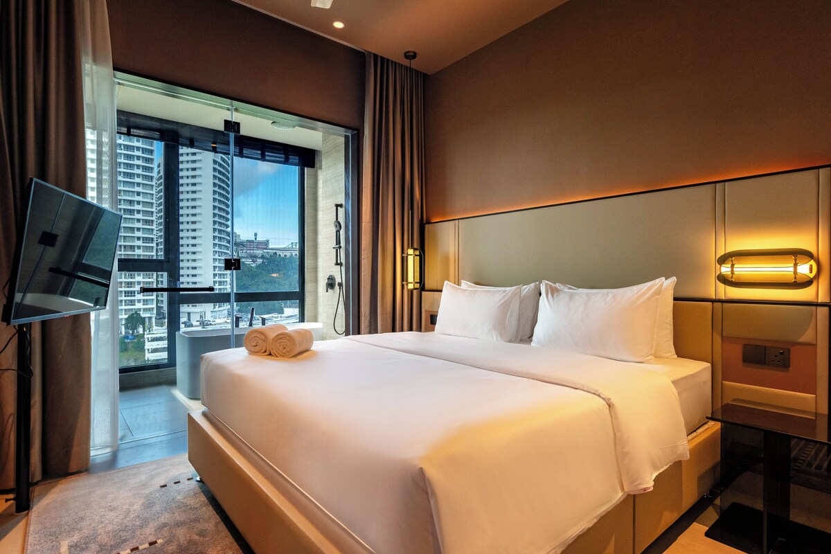 Habitación de hotel con una cama grande, televisor de pantalla plana, vista de la ciudad a través de un ventanal amplio y un balcón.