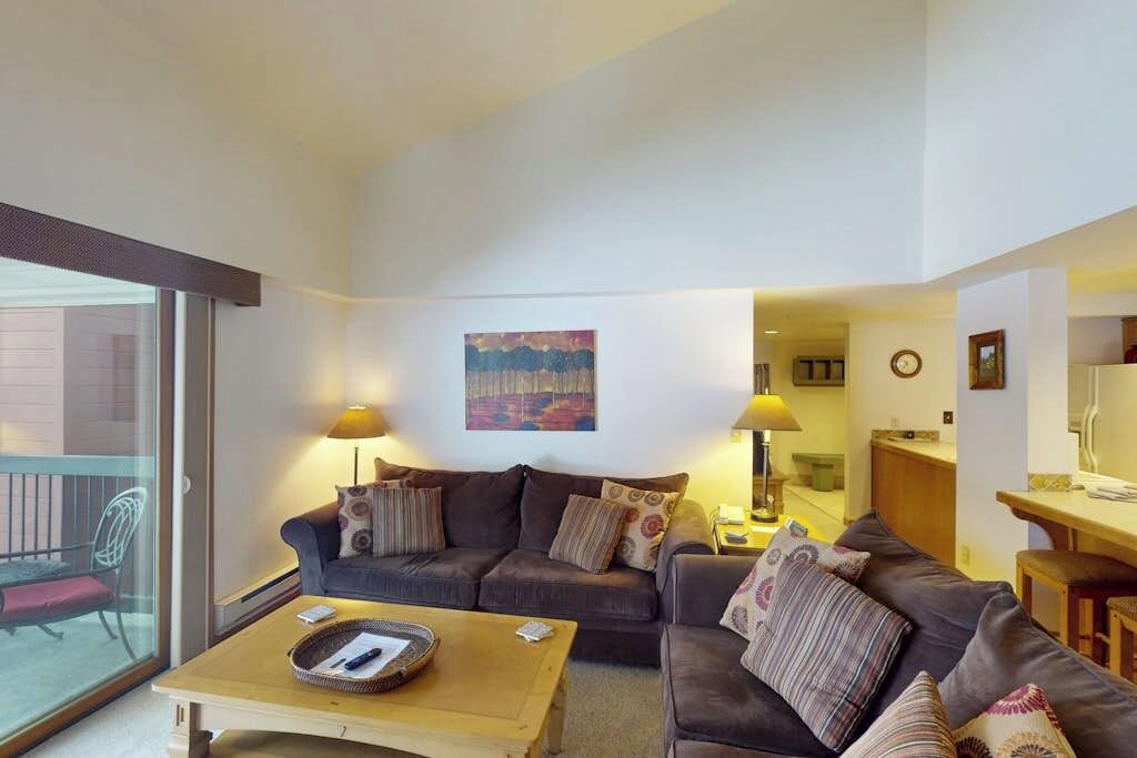 Uma sala de estar com um sofá, uma mesa de centro e duas luminárias. Há um quadro na parede e uma janela com vista.