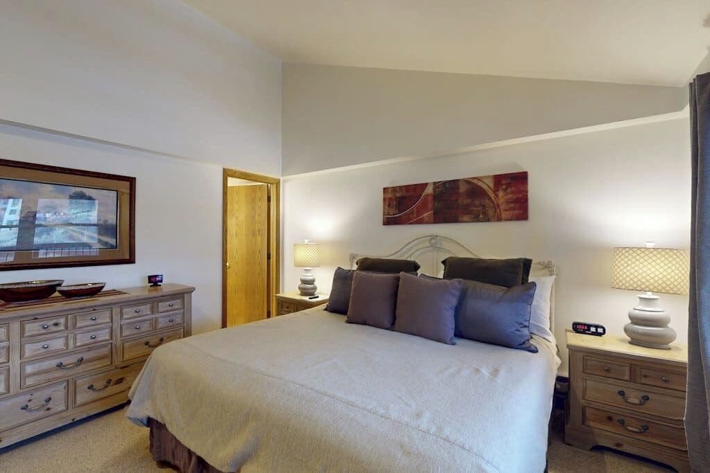 A bedroom with a bed, a dresser, a nightstand with a lamp, a painting on the wall, and a door.