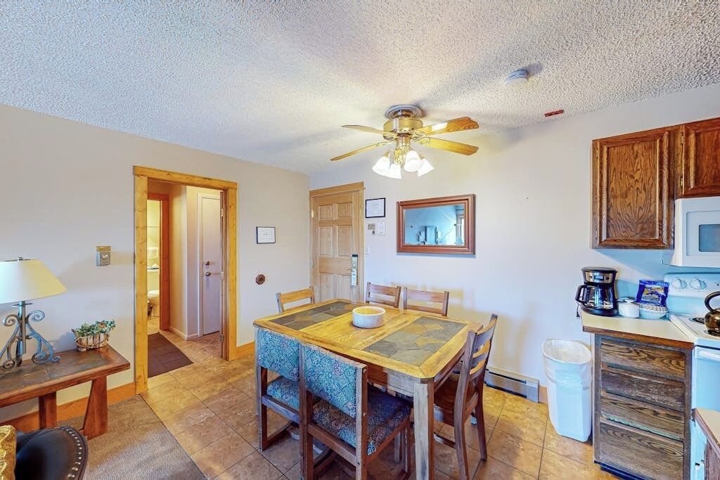 A dining area with a wooden table and chairs, a ceiling fan, a kitchen with a microwave, and a trash bin.