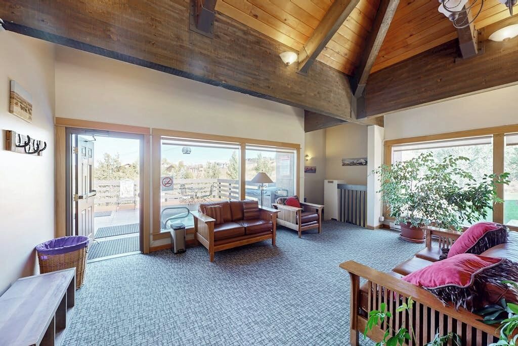 A spacious living area with a wooden ceiling, large windows, a sofa, a chair, and a potted plant.