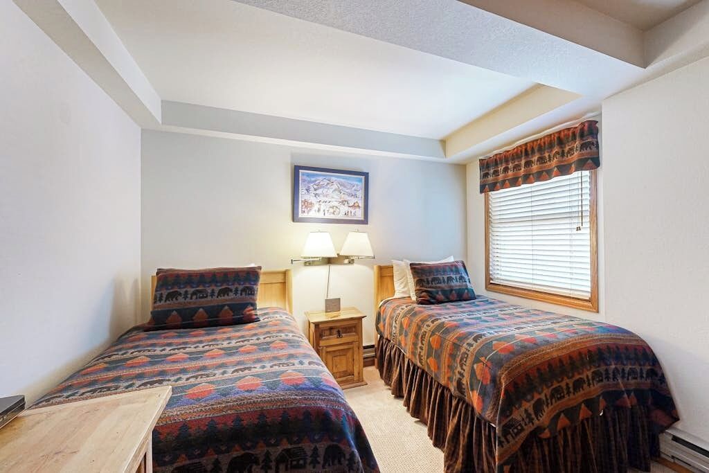 A room with two beds, a nightstand, a lamp, a framed picture on the wall, and a window with blinds.