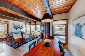 A spacious living area with a wooden ceiling, a dining table, and a large window offering a view of the outside. - Slopeside condo with pool, hot tubs, ski shuttle, fireplace, & deck (Steamboat Springs)
