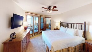 Ein Schlafzimmer mit einem Bett, einem Deckenventilator, einem Fernseher, einer Kommode und einer Glastür, die auf einen Balkon führt.