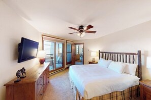 Ein Schlafzimmer mit einem Bett, einem Deckenventilator, einem Fernseher, einer Kommode und einer Glastür, die auf einen Balkon führt.
