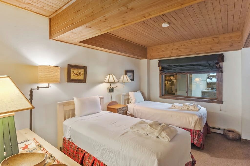 A room with two beds, a wooden ceiling, a window with a view, and a lamp.