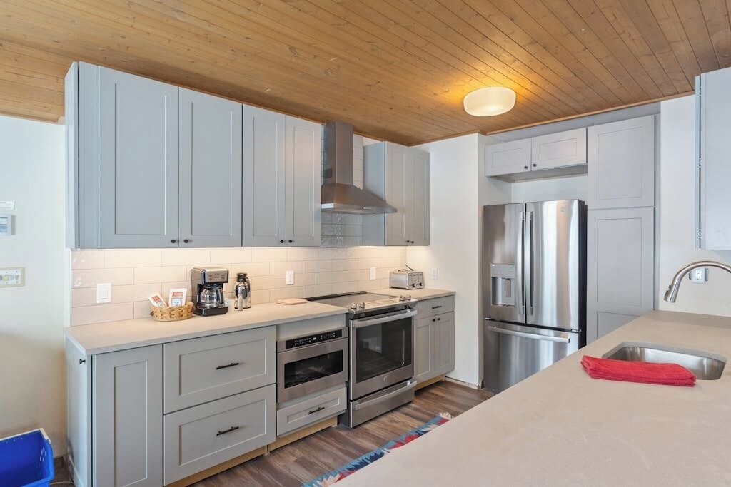 A modern kitchen with grey cabinets, stainless steel appliances, and a wooden ceiling.