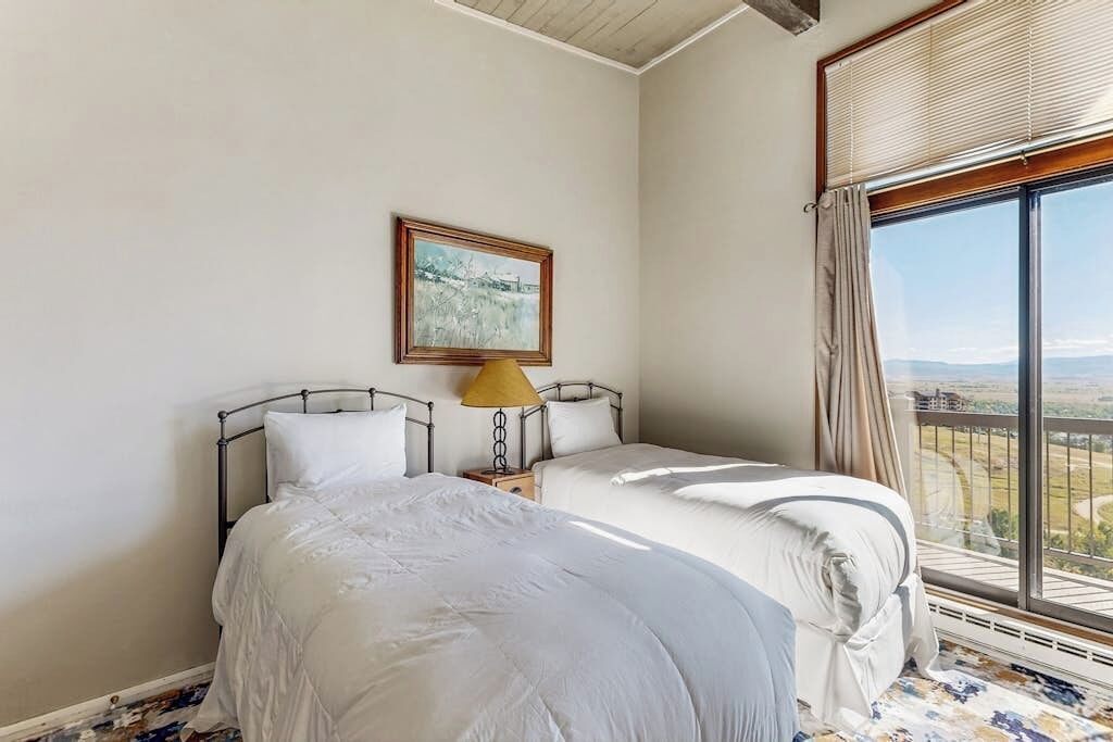 A bedroom with two beds, a nightstand, a lamp, a framed picture, and a large window with a view.