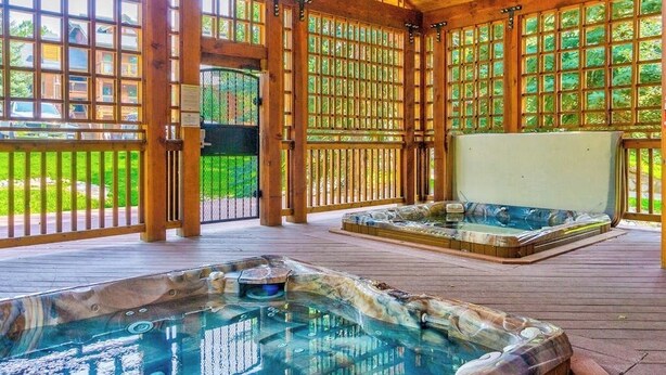 En træterrasse med jacuzzi og siddeplads, omgivet af store vinduer og udsigt til grøn natur.