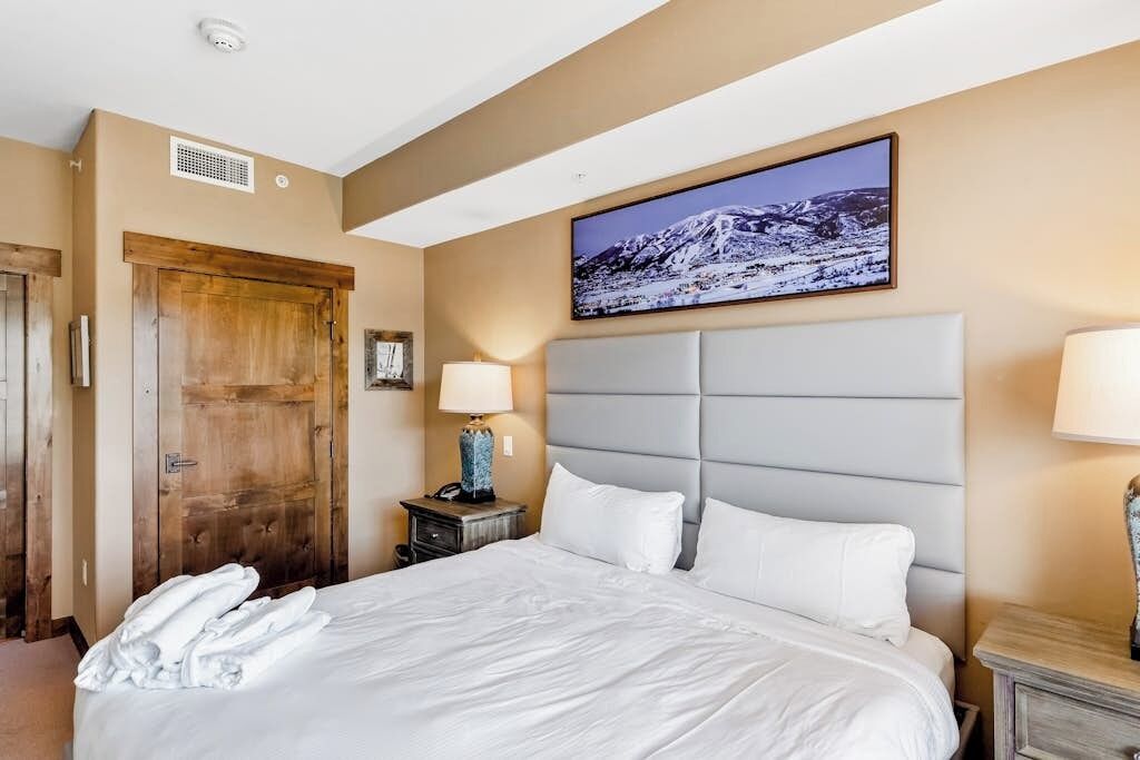 Une chambre d’hôtel avec un grand lit, une porte en bois, un tableau accroché au mur et une table de chevet avec une lampe.