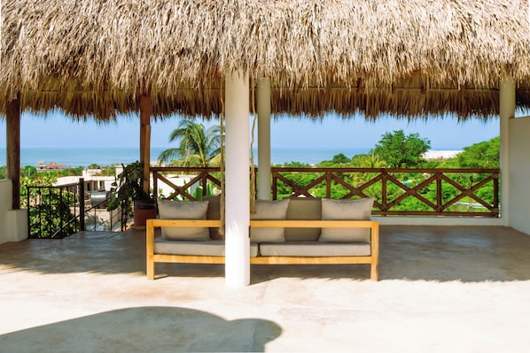 A covered outdoor seating area with a thatched roof, a cushioned bench, and a view of the ocean. - Ocean-view Villa with private Pool oasis (Puerto Escondido)