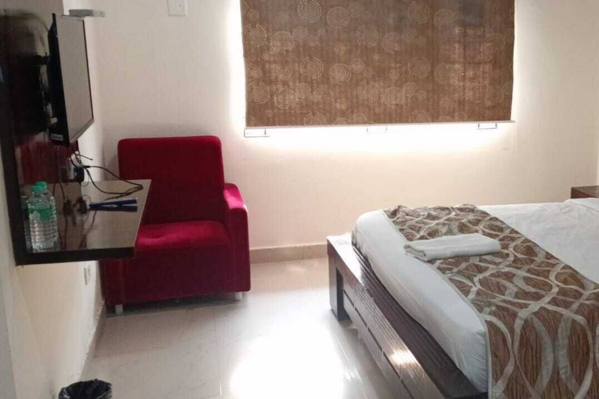 A bedroom with a bed, a red chair, a window with curtains, and a wall-mounted TV.