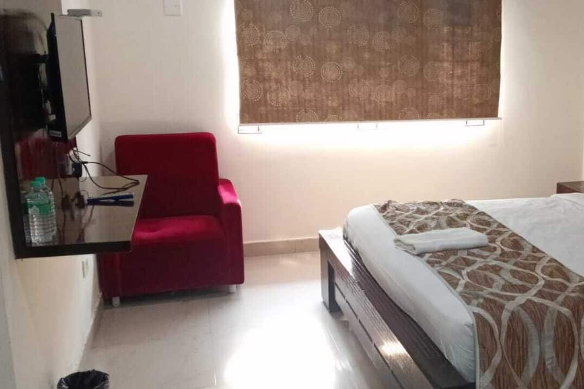 A bedroom with a bed, a red chair, a window with curtains, and a wall-mounted TV.