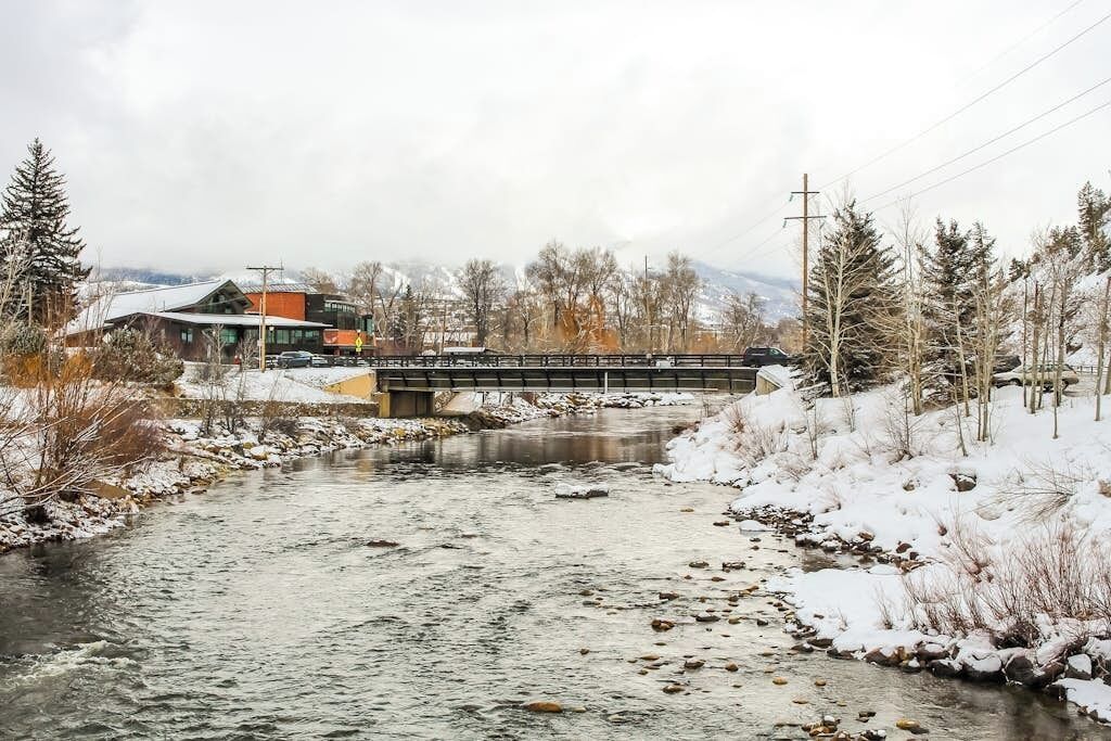 Un río fluye bajo un puente, con un edificio a la izquierda y árboles cubiertos de nieve a ambos lados.