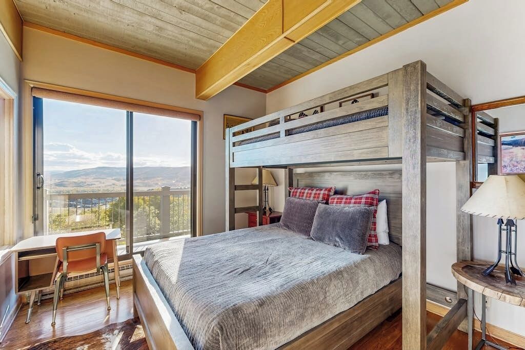 A room with a bunk bed, a desk, and a chair, with a view of the outdoors.