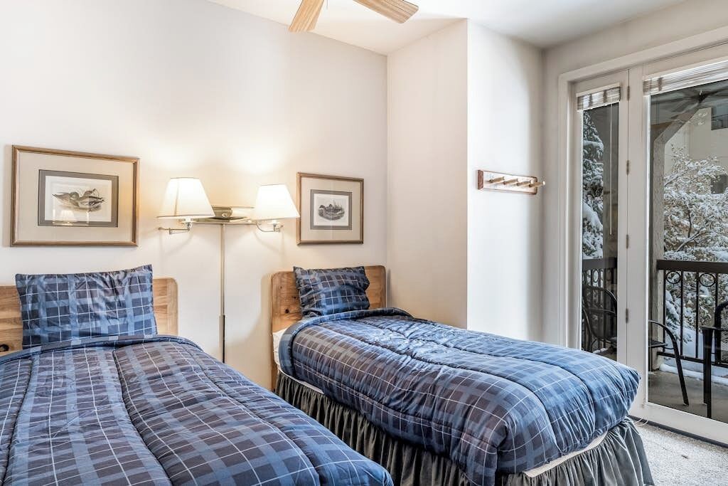 A room with two beds, a balcony with a view of snow-covered trees, and wall-mounted lamps.