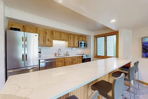 A modern kitchen with wooden cabinets, a stainless steel refrigerator, a microwave, and a large marble countertop. - 2nd-Floor Condo w/Mountain Views, Private Balcony, Fast WiFi, & Gas Fireplace (Steamboat Springs)