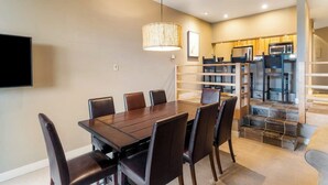A modern dining area with a wooden table and chairs, a hanging light fixture, and a kitchen in the background. - Two-story condo with ski view, pool, hot tub & gas fireplace - ski/walk to lifts (Steamboat Springs)