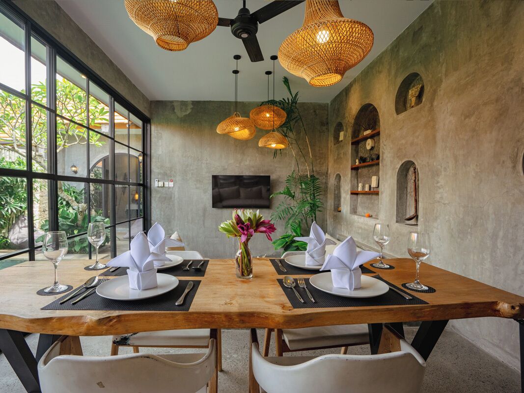 A dining area with a wooden table set for two, surrounded by white chairs. There are wicker pendant lights, a flat-screen TV mounted on the wall, and a potted plant on the table.