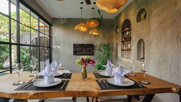 A dining area with a wooden table set for two, surrounded by white chairs. There are wicker pendant lights, a flat-screen TV mounted on the wall, and a potted plant on the table.