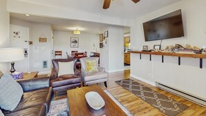 Uma sala de estar com ventilador de teto, TV de tela plana, espaço de jantar com cadeiras e cozinha com bancada.