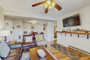 A living room with a ceiling fan, a flat-screen TV, a dining area with chairs, and a kitchen area with a countertop. - Lovely condo with shared pool, sauna, hot tub, tennis, free WiFi, gas fireplace (Steamboat Springs)