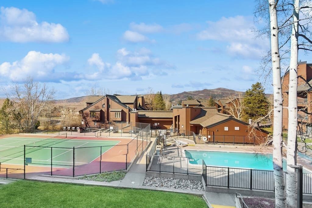 A residential area with a tennis court, a swimming pool, and houses with a hilly landscape in the background.