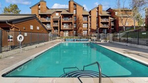 Piscina com deck de madeira, cercada por uma grade, e um edifício com várias varandas ao fundo.