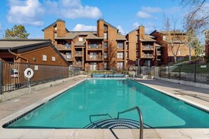 A swimming pool with a wooden deck, surrounded by a fence, and a building with multiple balconies in the background. - Lovely condo with shared pool, sauna, hot tub, tennis, free WiFi, gas fireplace (Steamboat Springs)