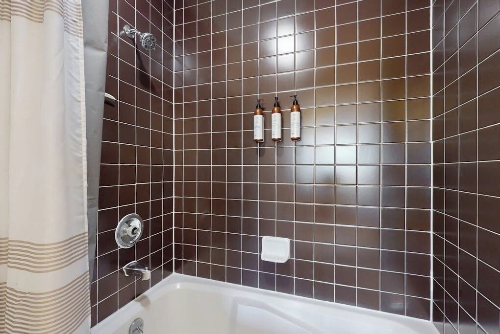 Une salle de bain avec une baignoire, des murs recouverts de carreaux de couleur marron foncé, un rideau de douche et trois bouteilles posées sur le mur.
