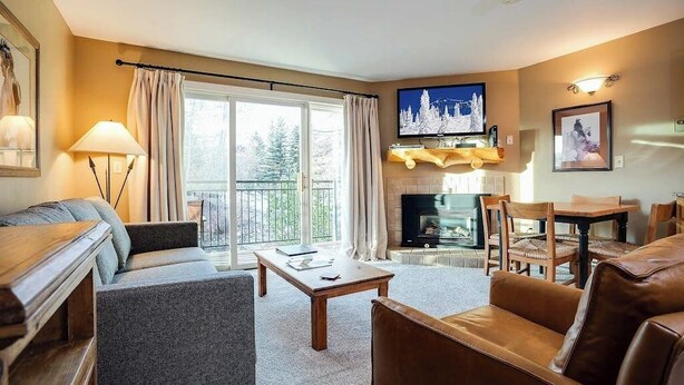 Sala de estar con sofá, sillón, mesa de centro, chimenea, televisor y balcón con cortinas.