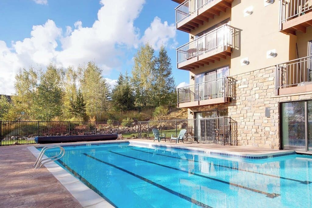 A swimming pool with a stone facade building, outdoor seating, and trees in the background.