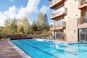 Una piscina con un edificio de fachada de piedra, mobiliario exterior y árboles al fondo.