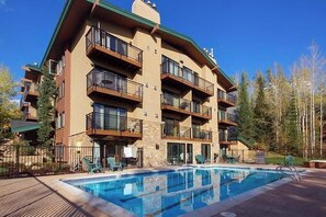 A multi-story hotel with balconies, a swimming pool, and a forested backdrop.