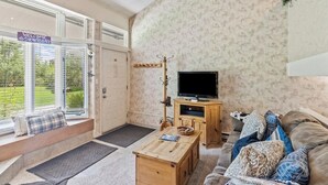 Ein gemütliches Wohnzimmer mit Fernseher, einem Holztisch, einer Couch mit gemusterten Kissen, einer Fensterbank mit freundlicher Willkommensanzeige und einer zur Außenseite führenden Tür.