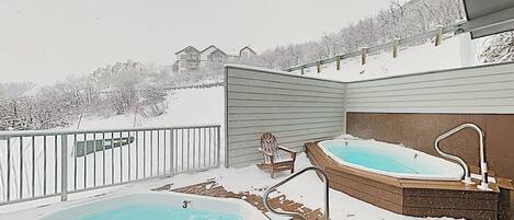 Ett bubbelbad och en pool på ett snötäckt däck med utsikt över hus i bakgrunden.