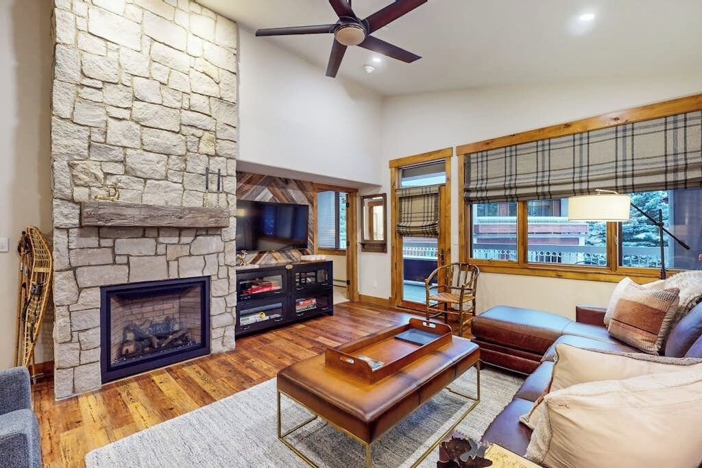 Una sala de estar con chimenea de piedra, ventilador de techo, sofá y ventanas amplias.