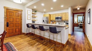A modern kitchen with wooden cabinets, a central island with bar stools, and a visible door leading to another room. - Spacious newly renovated condo w/shared pools, hot tubs, & ski shuttle (Steamboat Springs)