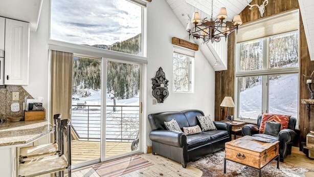 A spacious living area with a large window, a black leather sofa, a wooden coffee table, and a rustic chandelier. - Charming chalet with fireplace, loft & a true ski-in/out location (Telluride)