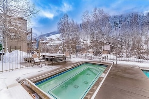 Ein Schwimmbecken mit Holzterrasse, umgeben von schneebedeckten Bäumen und Gebäuden.