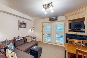 Ein Wohnzimmer mit einer Couch, einem Hocker, einem Holzesstisch, Stühlen, einem Fernseher und einem Kamin.