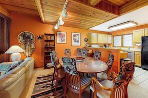 A cozy living room with a sofa, a dining table with patterned chairs, a kitchen with wooden cabinets, and a ceiling with exposed beams.