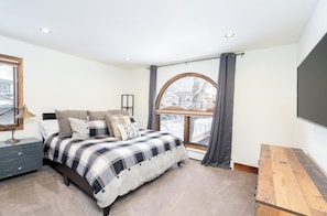 Ein Schlafzimmer mit Bett, Nachttisch, Schreibtisch und einem großen Fenster mit Vorhängen.