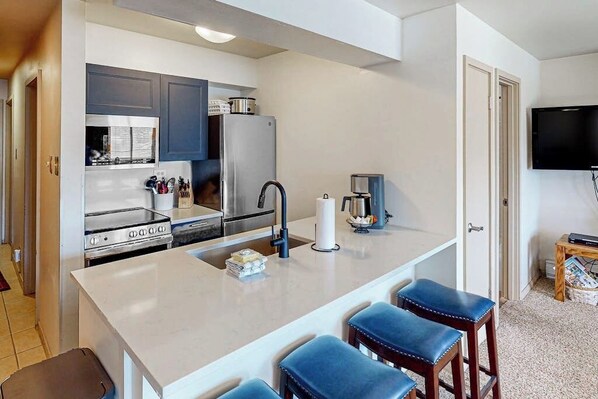 A compact kitchen with a central island, stainless steel appliances, and a flat-screen TV mounted on the wall. - Mountain getaway with modern comforts- pool, hot tub & summer activities (Steamboat Springs)