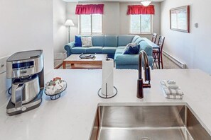 A modern kitchen with a stainless steel sink, a coffee maker, and a dining area with a blue sofa and red curtains. - Mountain getaway with modern comforts- pool, hot tub & summer activities (Steamboat Springs)