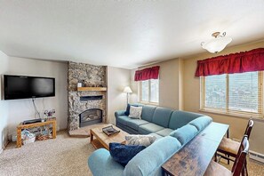 A living room with a stone fireplace, a flat-screen TV, a sofa, a coffee table, and a window with red valance. - Mountain getaway with modern comforts- pool, hot tub & summer activities (Steamboat Springs)