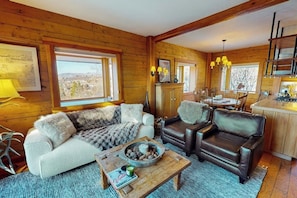 Ein gemütliches Blockhaus-Interieur mit Sofa, Sesseln und Essbereich, mit Holzwänden und großen Fenstern.