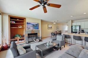A modern living room with a fireplace, a ceiling fan, and a kitchen area with bar stools. - Vail-View Condo w/ Balcony, BBQ, Large Indoor Pool-Near Skiing (Vail)