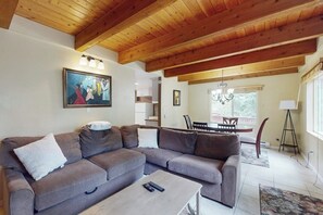 Ein Wohnzimmer mit Ecksofa, Couchtisch, einem Gemälde an der Wand und einem Essbereich mit einer Deckenleuchte.