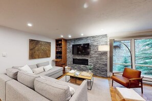 Ein modernes Wohnzimmer mit einem grauen Sofa, einer Holzregal, einem Flachbildfernseher an einer Steinwand und einem großen Fenster mit Blick auf Bäume.